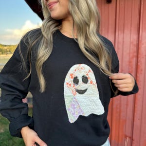 May include: A black long-sleeve crewneck sweatshirt featuring a large, central ghost-shaped applique. The ghost is constructed from quilted fabric patches in white, orange floral, and purple patterns, with black fabric eyes and a smiling mouth.