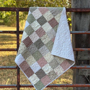 May include: A patchwork quilt featuring a grid of square fabric patches in muted shades of sage green, dusty rose, and cream. The quilt displays various subtle floral and geometric patterns on the squares and has a solid white backing.