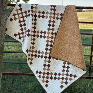 May include: A handmade quilt featuring a white background with a repeating chain pattern made of small brown, tan, and rust-colored squares. The quilt has a solid tan backing with a subtle mottled texture and intricate swirling machine quilting.