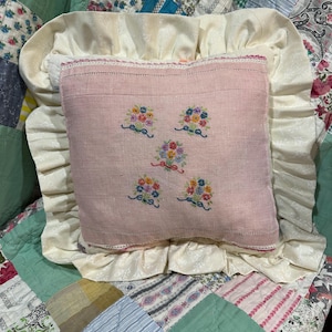 May include: A square pink linen throw pillow featuring five embroidered floral bouquet motifs in the center. The pillow is framed by a wide, cream-colored ruffled border with a subtle tonal pattern and a delicate pink lace trim edge.