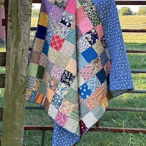 May include: A patchwork quilt constructed from numerous square fabric patches featuring a variety of floral, plaid, striped, and polka dot patterns in colors including pink, blue, yellow, green, and red, with a solid blue floral backing.