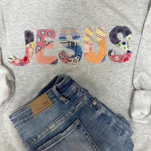 May include: A gray sweatshirt with the word "JESUS" appliqued in colorful patchwork fabric. Below the sweatshirt are folded blue denim jeans with a brown leather patch.