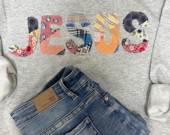 Puede incluir: Una sudadera gris con la palabra "JESUS" aplicada en tela de patchwork colorida. Debajo, unos vaqueros azules doblados con un parche de cuero marrón.