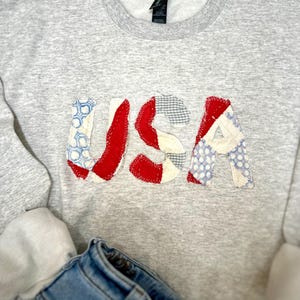 May include: A gray sweatshirt with the word "USA" appliqued across the front. The letters are made of red, white, and blue patterned fabric, resembling the American flag. A pair of blue jeans is visible in the lower portion of the image.