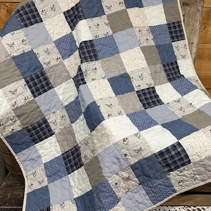 May include: A patchwork quilt in shades of blue, gray, and white. The quilt features a variety of patterns, including plaid, polka dots, and a chicken print. The quilt is made of soft fabric and is likely a handmade item.