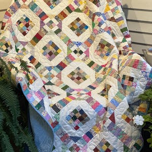 May include: A handmade quilt with a white background and colorful patchwork squares. The quilt features a repeating diamond pattern with various fabric patterns in shades of pink, blue, green, yellow, and purple. The quilt is draped over a chair.