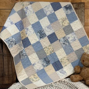 May include: A patchwork quilt featuring squares in shades of blue, cream, and tan. The squares display various patterns, including floral prints, polka dots, and plaid. The quilt is likely handmade, with visible stitching and a soft, quilted texture. A cozy, decorative item.