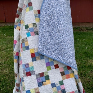May include: A patchwork quilt with a white background and squares of various colors and patterns. The quilt is folded in half, revealing a blue floral patterned backing.