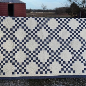 May include: A white and navy blue quilt with a checkered pattern. The quilt is hanging on a fence in front of a field.