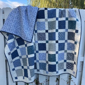 May include: A handmade quilt with a patchwork design in various shades of blue and white. The quilt features square blocks with different patterns, including floral and geometric prints. The quilt is draped over a white fence, showcasing its intricate details.