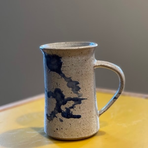 Peut inclure: Une tasse en céramique tachetée avec une anse. La tasse est beige avec des éclaboussures de peinture bleu foncé. La tasse est posée sur une surface jaune.