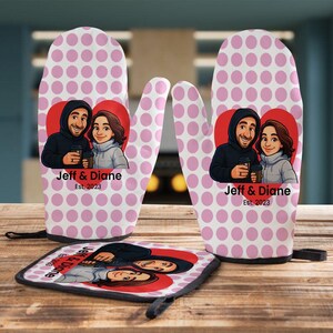 May include: A set of kitchen accessories featuring a pink polka dot pattern. The set includes two oven mitts and a pot holder. Each item has a red heart with a cartoon couple and the text "Jeff & Diane Est. 2023".
