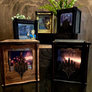 May include: Wooden display boxes with Magic: The Gathering cards. The boxes are stained in various shades of brown and black. Each box features a decorative emblem. A small Fallout pin is attached to one box. A small plant is visible.