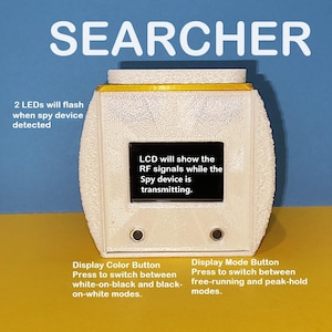 Könnte beinhalten: Ein weißes Gerät mit gelbem Rand, beschriftet mit "SEARCHER". Das Gerät hat einen kleinen LCD-Bildschirm, der "LCD will show the RF signals while the Spy device is transmitting." anzeigt. Unter dem Bildschirm befinden sich zwei Knöpfe mit den Beschriftungen "Display Color Button" und "Display Mode Button".