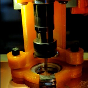 May include: Close-up of a drill bit attached to a metal spindle, surrounded by an orange plastic housing. The drill bit is positioned over a circular metal plate.