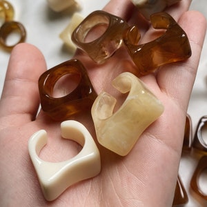 May include: A collection of resin rings in various shapes and colours, including brown, beige, and white. The rings are all unique and have a modern, minimalist aesthetic.