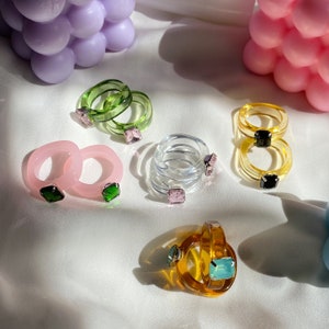 May include: Assortment of colourful clear plastic rings with various coloured gem-like stones. The rings are stacked and arranged on a white surface.
