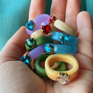 Puede incluir: Una pila de anillos de corazón coloridos con gemas de varios colores. Los anillos están hechos de plástico y tienen un acabado brillante.