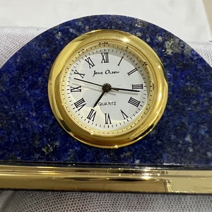 May include: A gold-toned clock with Roman numeral markings on a blue lapis lazuli base. The clock face reads "Jens Olsen Quartz".