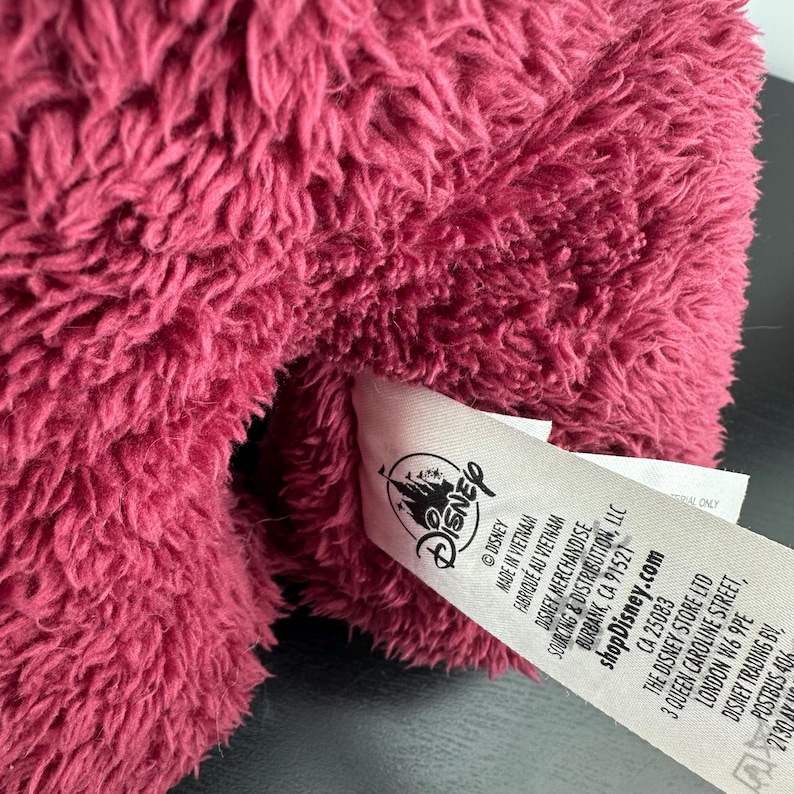May include: Close-up of a plush toy in a deep pink color. The toy's fur is textured and fluffy. A white tag with the Disney logo and text is attached to the toy.