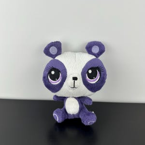May include: A plush panda toy featuring a white face with large purple patches around the eyes, purple ears, and a purple body with a white belly patch. The toy has embroidered facial features, including long eyelashes and a small black nose.