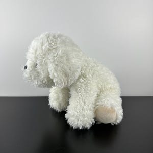 May include: A white plush dog toy with a fluffy, textured coat. The toy has black eyes and a light tan paw pad. The dog is sitting on its haunches, with its head turned to the left.