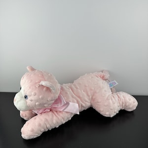 May include: A pink plush cat toy with a textured surface and a pink bow. The cat has blue eyes and white accents on its face. The toy is lying down on a black surface.