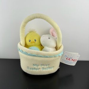 May include: A soft, cream-colored Easter basket with a yellow handle, containing a yellow chick and a white bunny. The basket has a light blue scalloped trim and the embroidered text "My First Easter Basket". A "Gund" tag is attached.