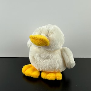 May include: White plush duck toy with yellow beak and feet. The duck is sitting on a black surface.