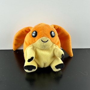 May include: Orange and yellow plush toy with black eyes and a smiling mouth. The toy has large, floppy ears and small, black feet.
