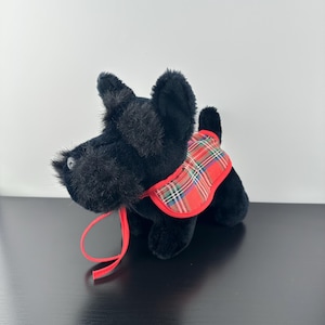 May include: A plush black Scottish Terrier dog with a red and green plaid vest. The dog has a red ribbon around its neck. The toy is sitting on a black surface.