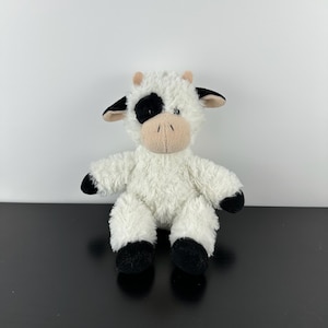 Puede incluir: Un peluche de vaca con cuerpo blanco, manchas negras y pezuñas negras. La vaca tiene un hocico y orejas beiges. El juguete está diseñado para sentarse erguido y tiene una textura suave y esponjosa. Es un animal de peluche.