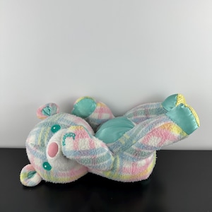 May include: A plush teddy bear in pastel colors, featuring a plaid pattern in pink, blue, and yellow. The bear has a pink nose, teal accents, and the word "Pleasur" embroidered on its chest. The bear is lying on its back.