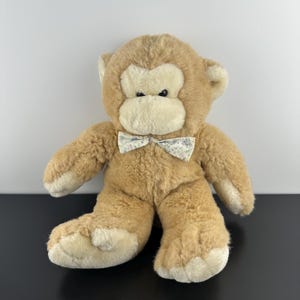 May include: A plush monkey toy with tan fur and a cream-colored face. The monkey features dark eyes, a rounded snout, and a small bow tie around its neck. The bow tie is made of a light-colored fabric with a delicate blue and pink floral pattern.