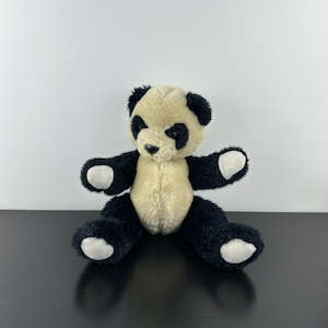 May include: A soft panda bear toy with a cream-coloured body, black arms, legs, and ears, and white accents on the paws. The stuffed animal is sitting with its arms outstretched, and its black eyes and nose are visible.