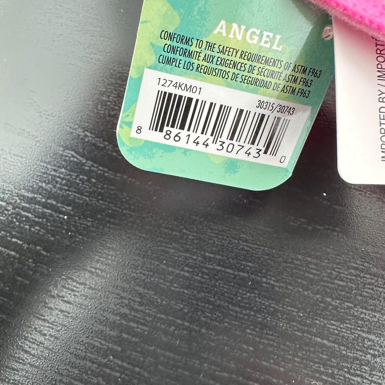 Op de afbeelding: Een close-up van een productlabel met het woord "ANGEL" bovenaan. Het label is groen en wit met een barcode en de tekst "CONFORMS TO THE SAFETY REQUIREMENTS OF ASTM F963".