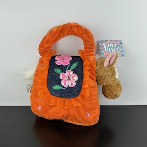 May include: An orange corduroy handbag with a denim pocket featuring embroidered pink flowers and green leaves. The bag has a handle and a small stuffed animal attached. The tag reads "Love to GO!"