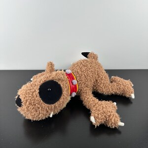 May include: A plush toy dog with brown fur, a red collar with grey accents, and a large black eye. The dog has white claws and black nose and ears. The toy is lying on a black surface.