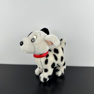 May include: A plush dalmatian dog toy with white fur and black spots. The stuffed animal features large, round, black and white eyes, a black nose, and a smooth, bright red plastic collar around its neck. It is standing in a side profile.