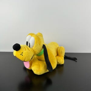 Puede incluir: Un juguete de peluche amarillo con forma de perro con orejas negras, nariz negra y lengua rosa. El perro tiene ojos blancos y un collar verde. El juguete está tumbado sobre una superficie negra.