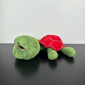 May include: A plush turtle toy with a green head and limbs, and a red shell. The turtle has large, expressive eyes and is lying on a dark surface. The toy is soft and cuddly, perfect for children.