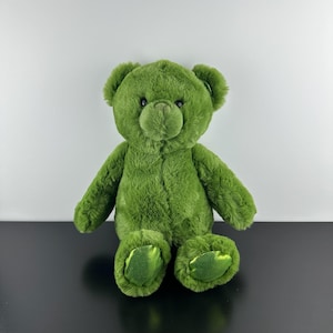 Könnte beinhalten: Ein Plüsch-Teddybär mit weichem, olivgrünem Fell. Der Bär hat runde Ohren, eine gestickte Nase und dunkle, runde Augen. Die Fußsohlen bestehen aus einem kontrastierenden, glänzenden, metallisch grünen Stoff, der das Licht reflektiert.