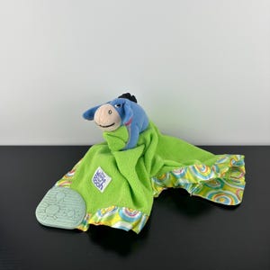 May include: A plush Eeyore toy from Winnie the Pooh, with a blue head and a green blanket. The blanket has a colorful, patterned border and a mint green teether. The Winnie the Pooh logo is visible on the blanket.