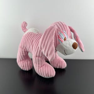 Puede incluir: Un juguete de peluche de perro rosa con textura de pana. El perro tiene orejas caídas, un hocico blanco y una nariz marrón. Los ojos están decorados con rayas de colores. La espalda del perro tiene un parche de lunares. Un peluche lindo y tierno.