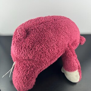 May include: A plush toy in a deep rose color, featuring a soft, textured exterior. The toy has a rounded body, small ears, and two legs with white-colored feet. A white string is attached to the toy.