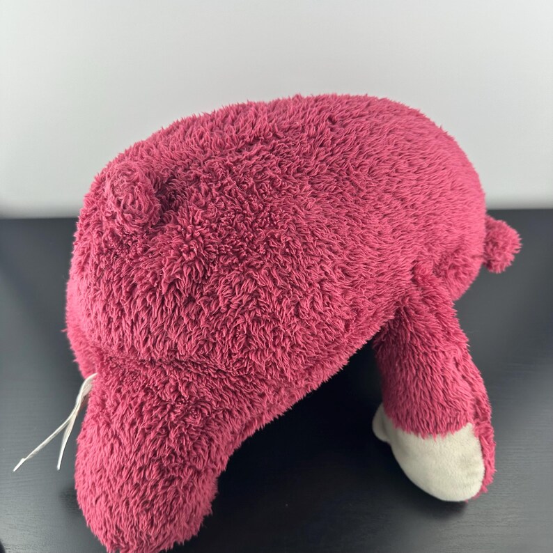 May include: A plush toy in a deep rose color, featuring a soft, textured exterior. The toy has a rounded body, small ears, and two legs with white-colored feet. A white string is attached to the toy.