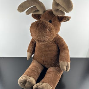 Peut inclure: Une peluche en forme d'élan avec une fourrure marron douce, de grands bois beiges et des yeux sombres. Ce jouet en peluche présente un museau arrondi, de petites oreilles et des sabots beiges sur les bras et les jambes, en position assise.
