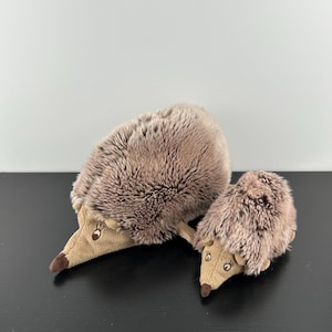 May include: Two plush hedgehog toys, one larger than the other, featuring soft, shaggy, light brown faux fur on their backs and smooth, tan-colored fabric on their faces, each with a small dark brown nose and embroidered eyes.