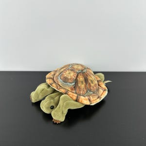 May include: A plush turtle toy with a detailed, brown and tan shell and olive green limbs. The toy is positioned on a black surface against a white background. The turtle's shell has a realistic pattern.