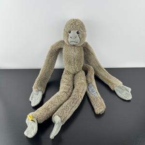May include: A soft toy monkey with long limbs. The monkey is mainly brown with grey hands and feet. It has a grey face and a small yellow tag attached to one foot.